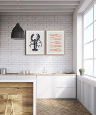 A white kitchen island with wooden panels and a wooden stool, and behind it a white brick wall with a blue lobster print and a pink fish print, with a wooden and white counter below it