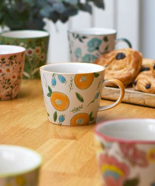 A close-up shot of a white mug with orange fruit illustrations on it, stood on a wooden counter with colorful floral mugs surrounding it and a wooden board with pastries on it behind it