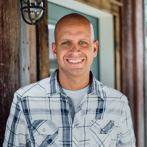 Image of a man in a white and blue plaid shirt standing in front of a wooden exterior.