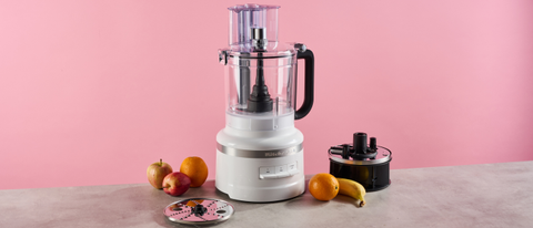 A white colored KitchenAid 13 cup food processor photographed against a pink background, sitting on a great stone-effect surface. The food processor is surrounded by apples, oranges and a banana. The accessory storage caddy is pictured to the right of the processor, and the reversible grating disc is on the left.