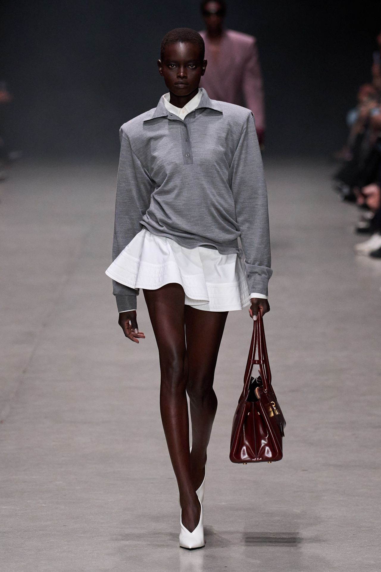 Layering staples for spring 2026 is shown in a photo from Stella McCartney's runway show, with a model walking while wearing a gray polo shirt over a white button-down shirt styled with a white ruffled mini skirt, white pointed pumps, and a structured burgundy red bag