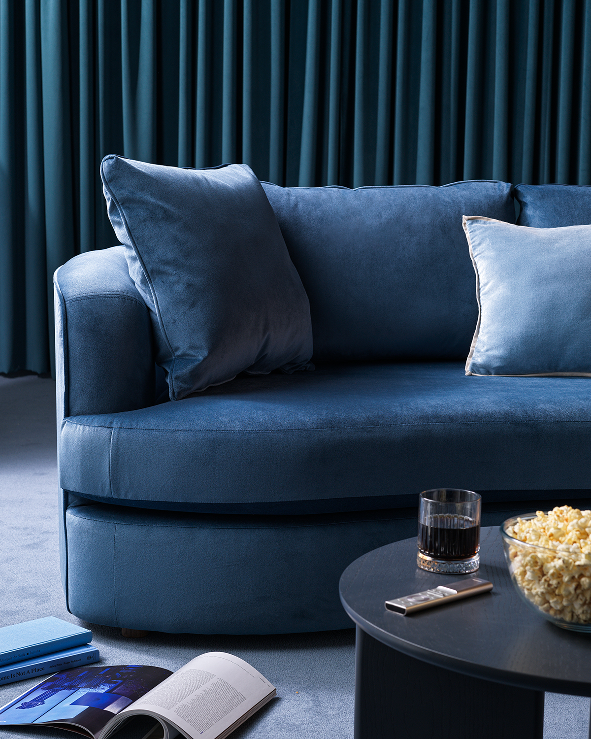 Close-up image of half of a blue velvet, curved sofa. The back wall is covered in dark blue curtains, and there is a round, navy coffee table in front of the sofa with a bowl of popcorn and a glass on it.