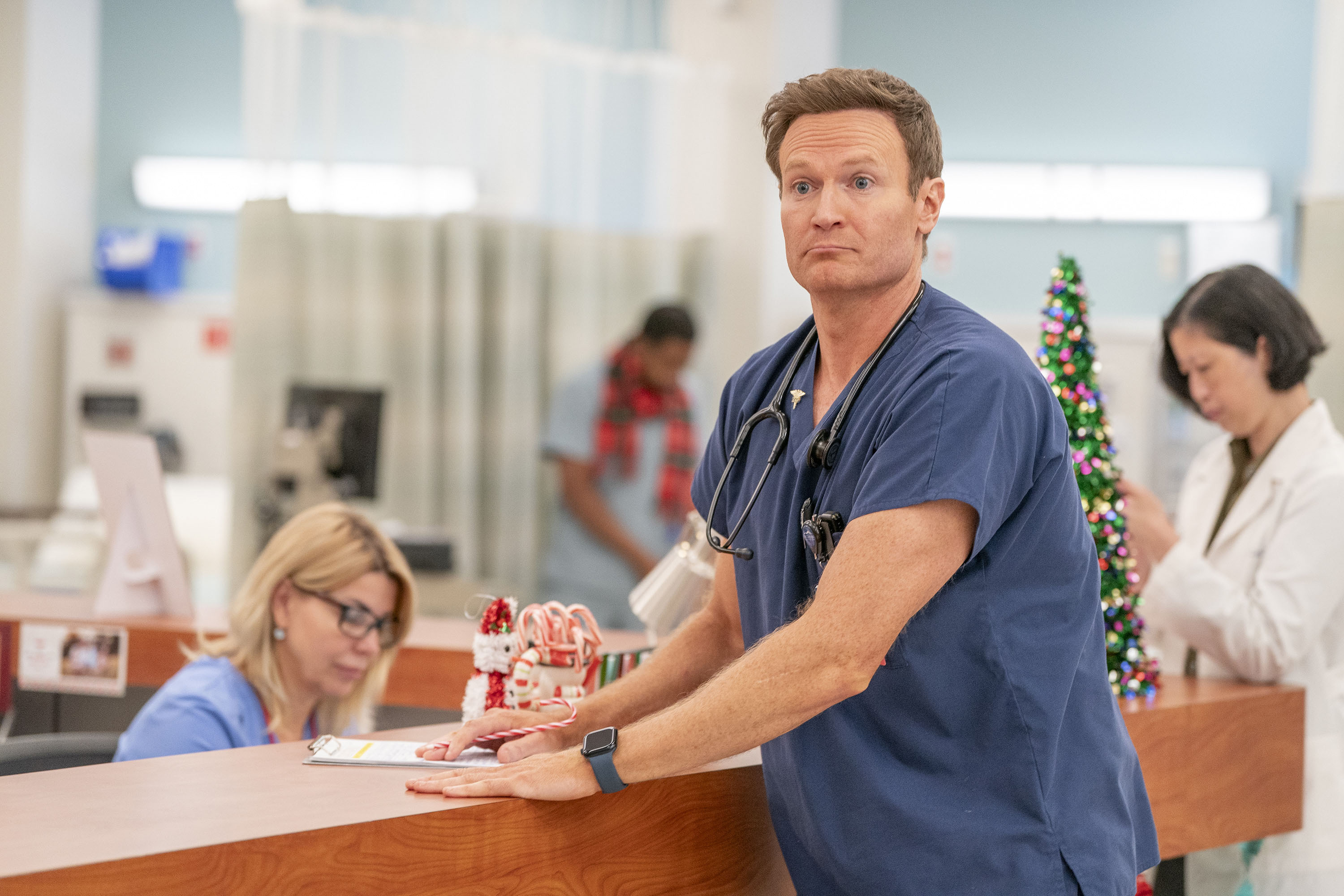 Josh Lawson as Dr Bruce, standing at reception in St. Denis Medical looking puzzled.