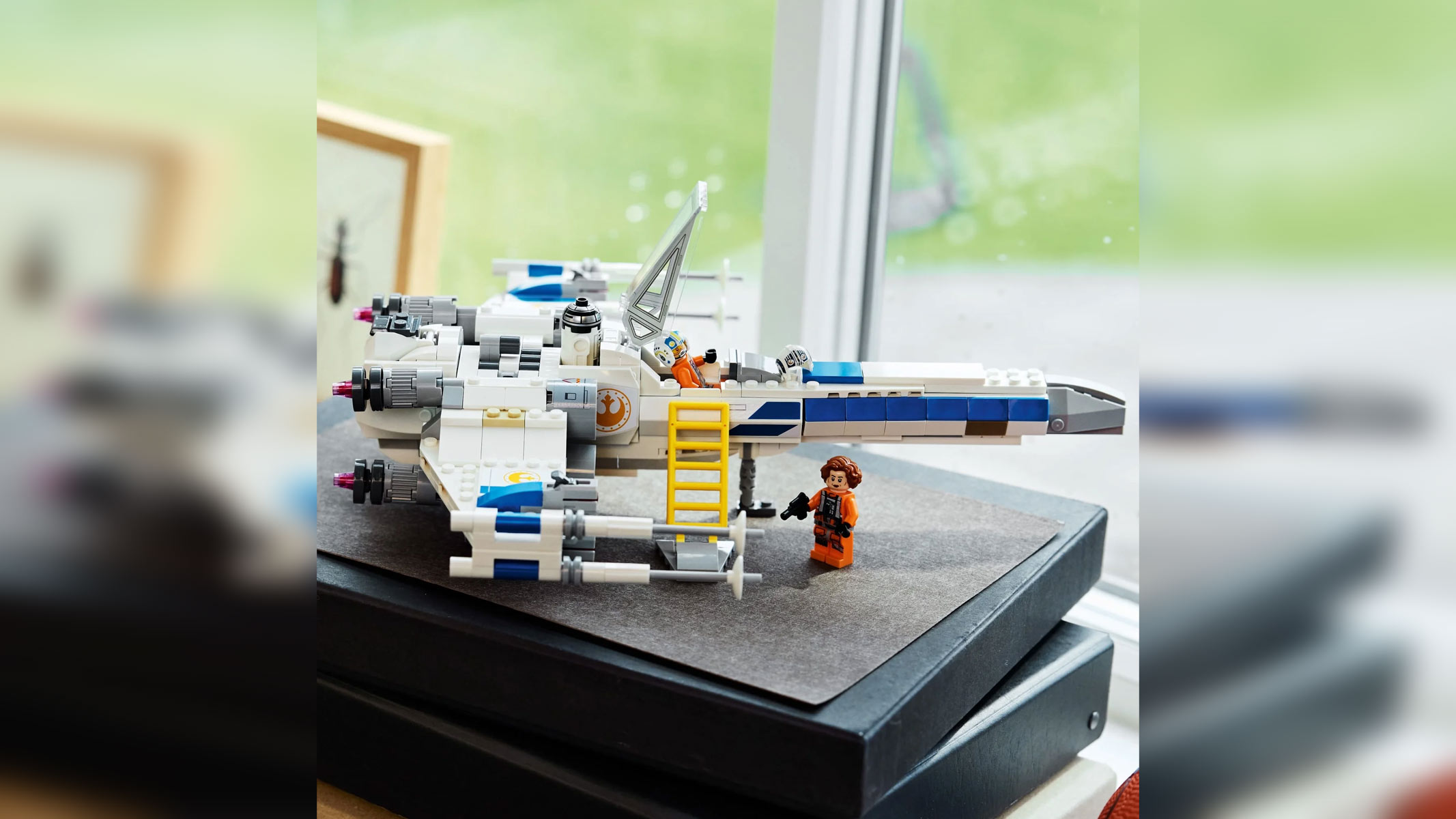 A white and blue spaceship is displayed on a in front of a window along with a yellow ladder and a minifigure dressed in orange.