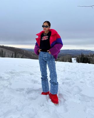 Woman standing in the snow and wearing a multicolor coat, jeans, and red shoes.