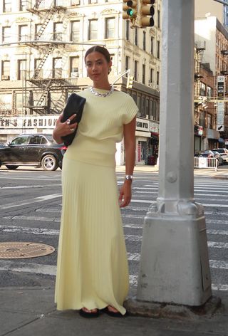 A woman wearing a silver necklace, silver bangle, black clutch bag, a yellow knit top with a matching maxi skirt and black flip flops.