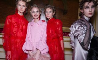 Four female models wearing looks from Simone Rocha's collection. Two models are wearing red semi-sheer dresses with sleeves and red detail. Another model is wearing a light pink belted coat with buttons. And the fourth model is wearing a grey high neck piece with sleeves and she is holding a black fur stole. All the models are wearing earrings