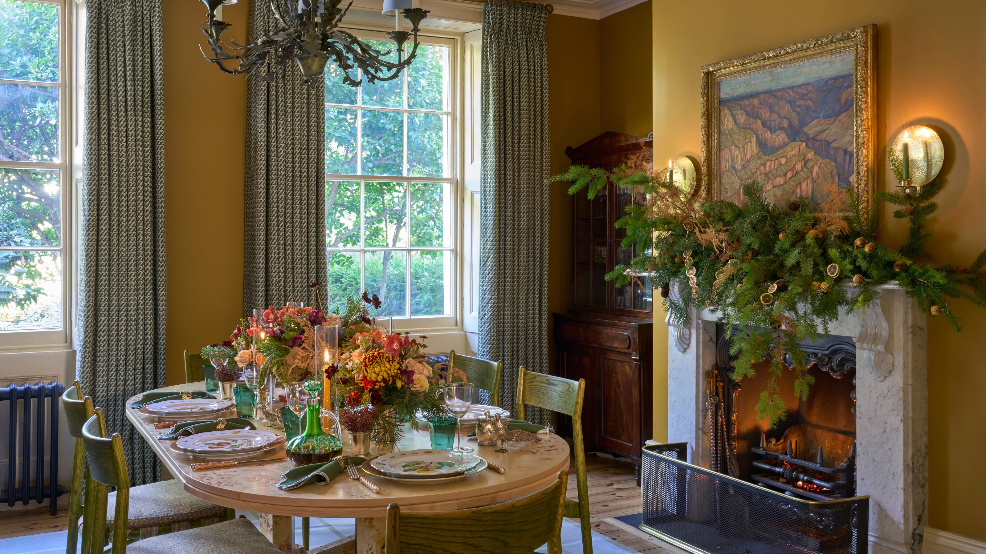 Elegant dining room featuring golden-yellow walls and windows with green and beige patterned curtains. A large wooden oval table is set with a colorful floral centerpiece. The seating consists of moss-green wooden chairs.