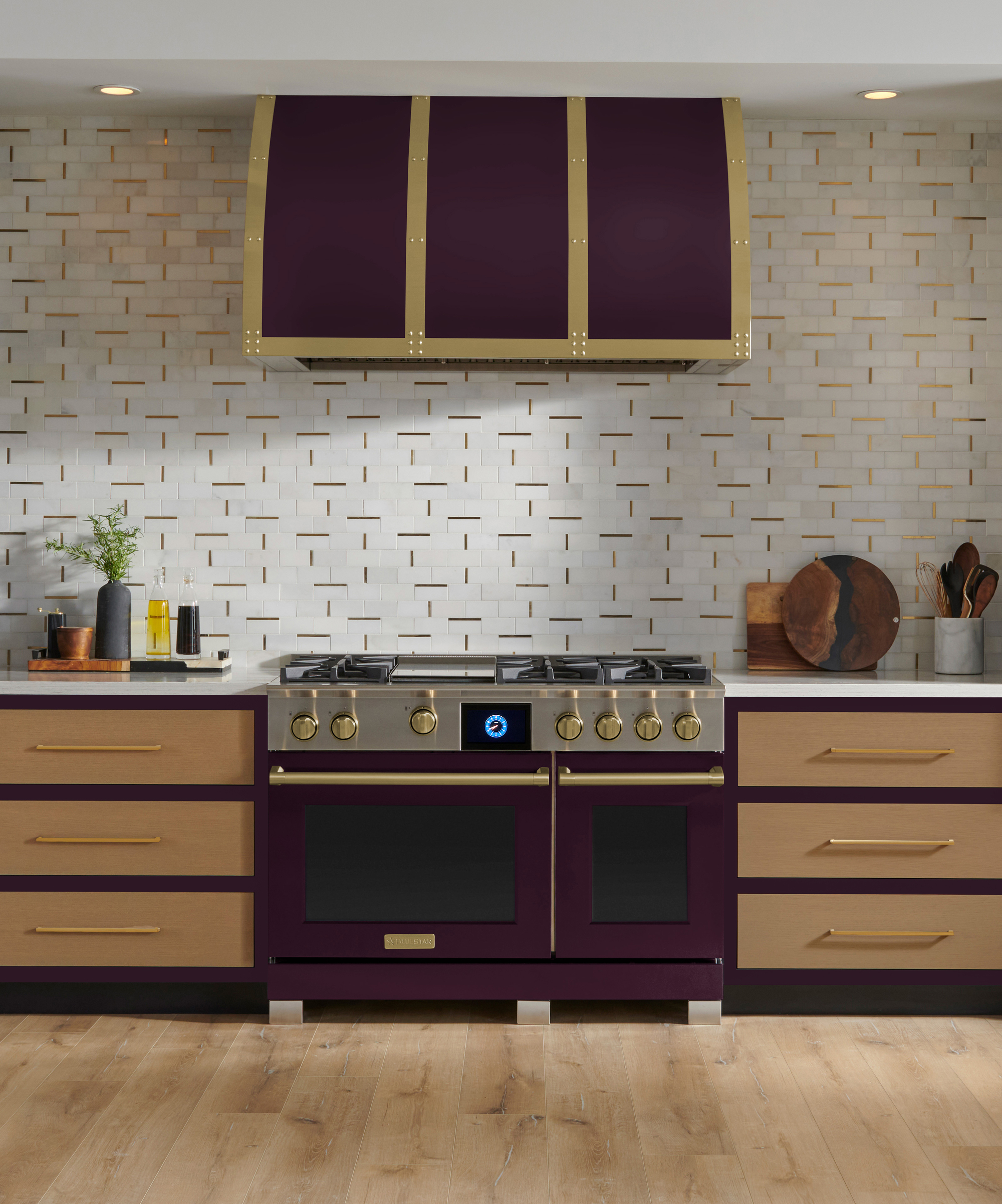 Image of a white kitchen with a purple oven and matching hood. The cabinetry is purple with wooden drawers, and the walls are white and gold subway tiles.