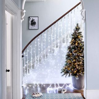 White staircase with hanging fairy lights next to a big green Christmas tree in a vase