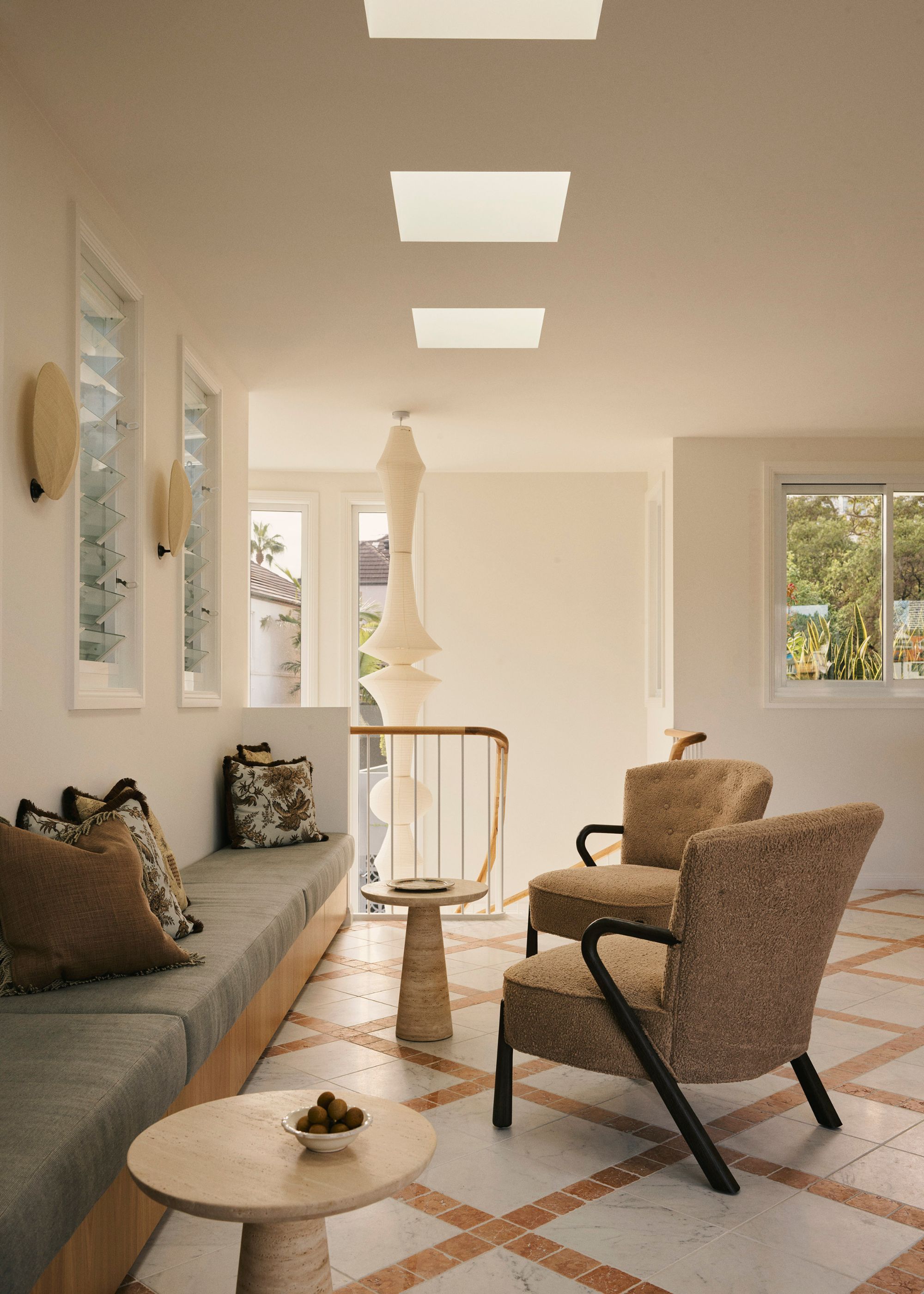 A living room with tiled flooring, a bench, patterned pillows, stone side tables, and a pair of sofa chairs, by a staircase with a big paper pendant