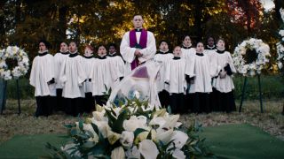 Burn Gorman oversees a funeral with a children's choir singing behind him in Beetlejuice Beetlejuice.