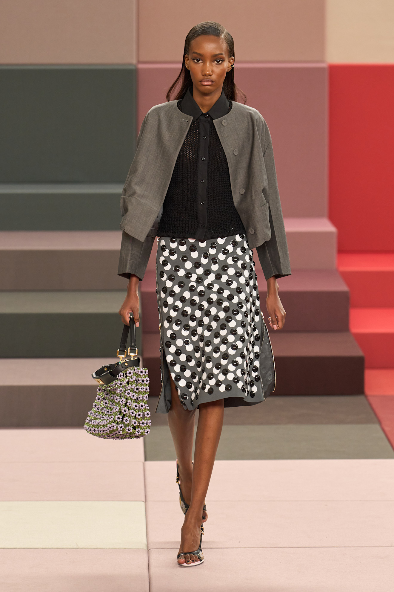 Layering staples for spring 2026 are shown in a photo from Fendi's runway show, with a model walking while wearing a black knit polo top layered under a gray blazer styled with a gray embellished pencil skirt, two-tone sandals, and a gray embellished bag