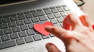 A finger touches a red paper heart on top of a laptop keyboard