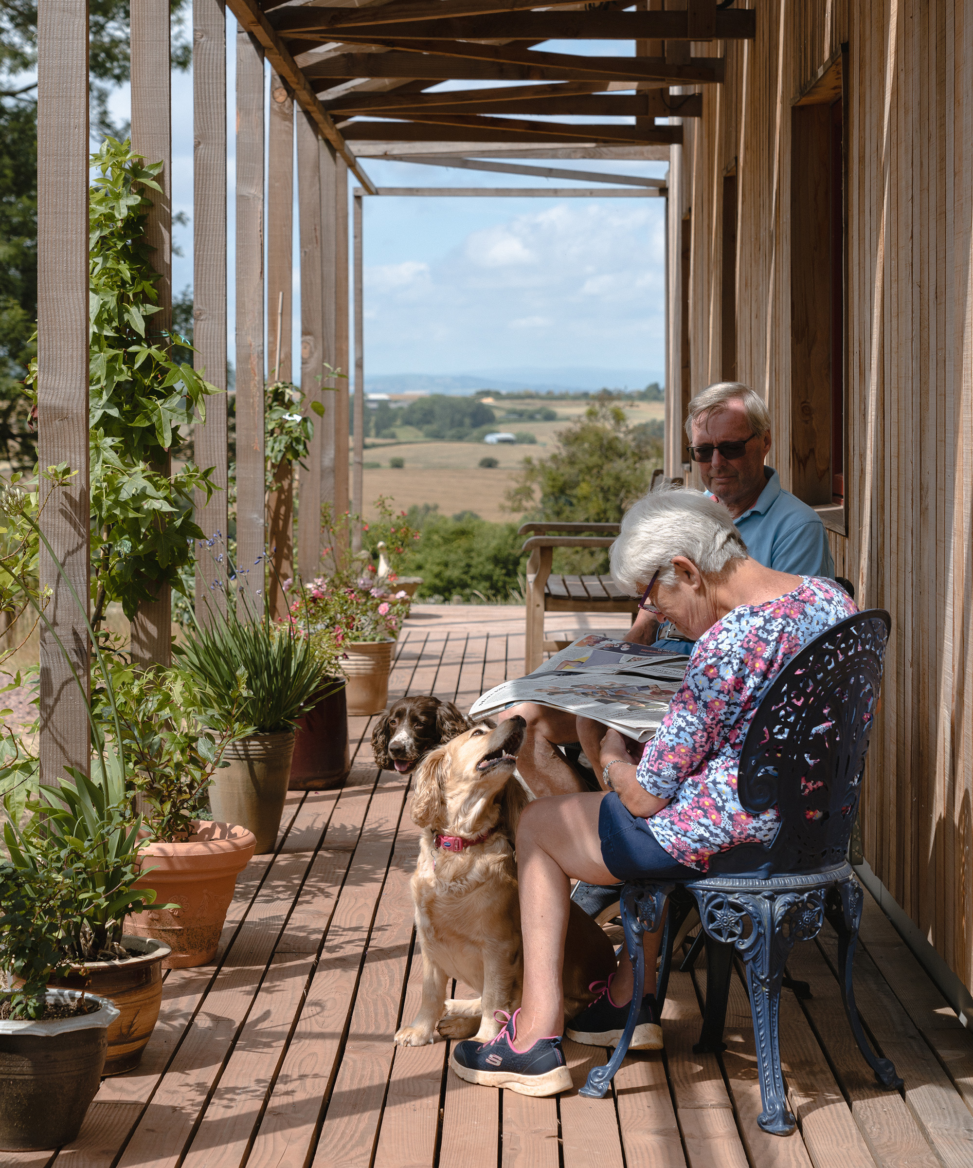 Elderly couple sits on a wooden veranda which is covered with a wooden pergola. They are relaxing and enjoying the surrounding countryside views with their dogs