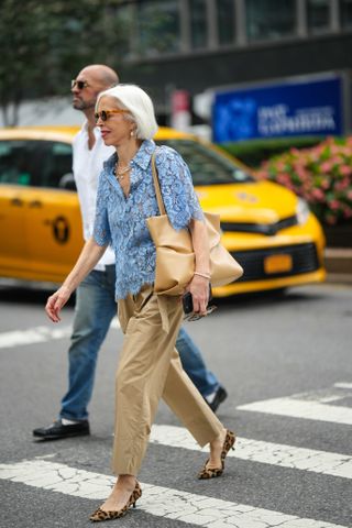 A woman wears sunglasses, a blue floral embroidery shirt, a beige leather bag, beige pants, brown kitten heels leopard print pointed shoes, outside Proenza Schouler, during New York Fashion Week