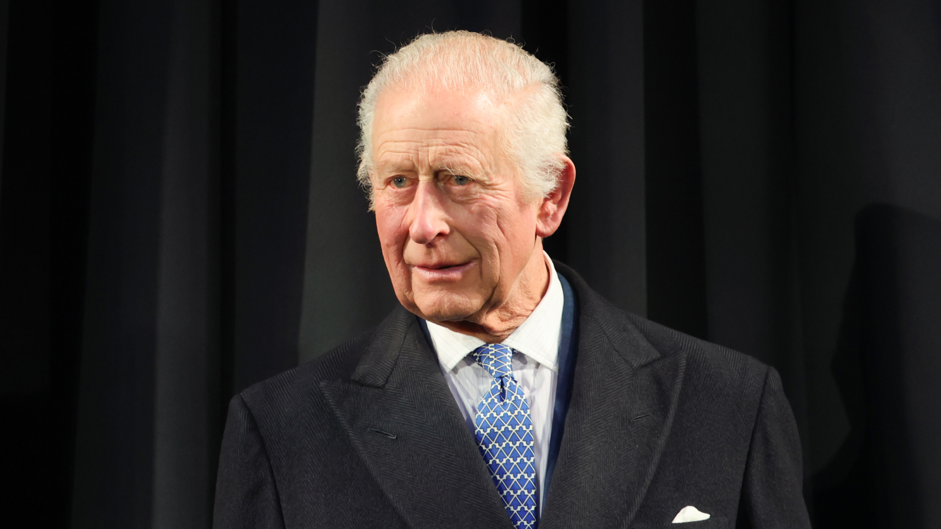 King Charles wears a dark overcoat with a white shirt and blue patterned tie