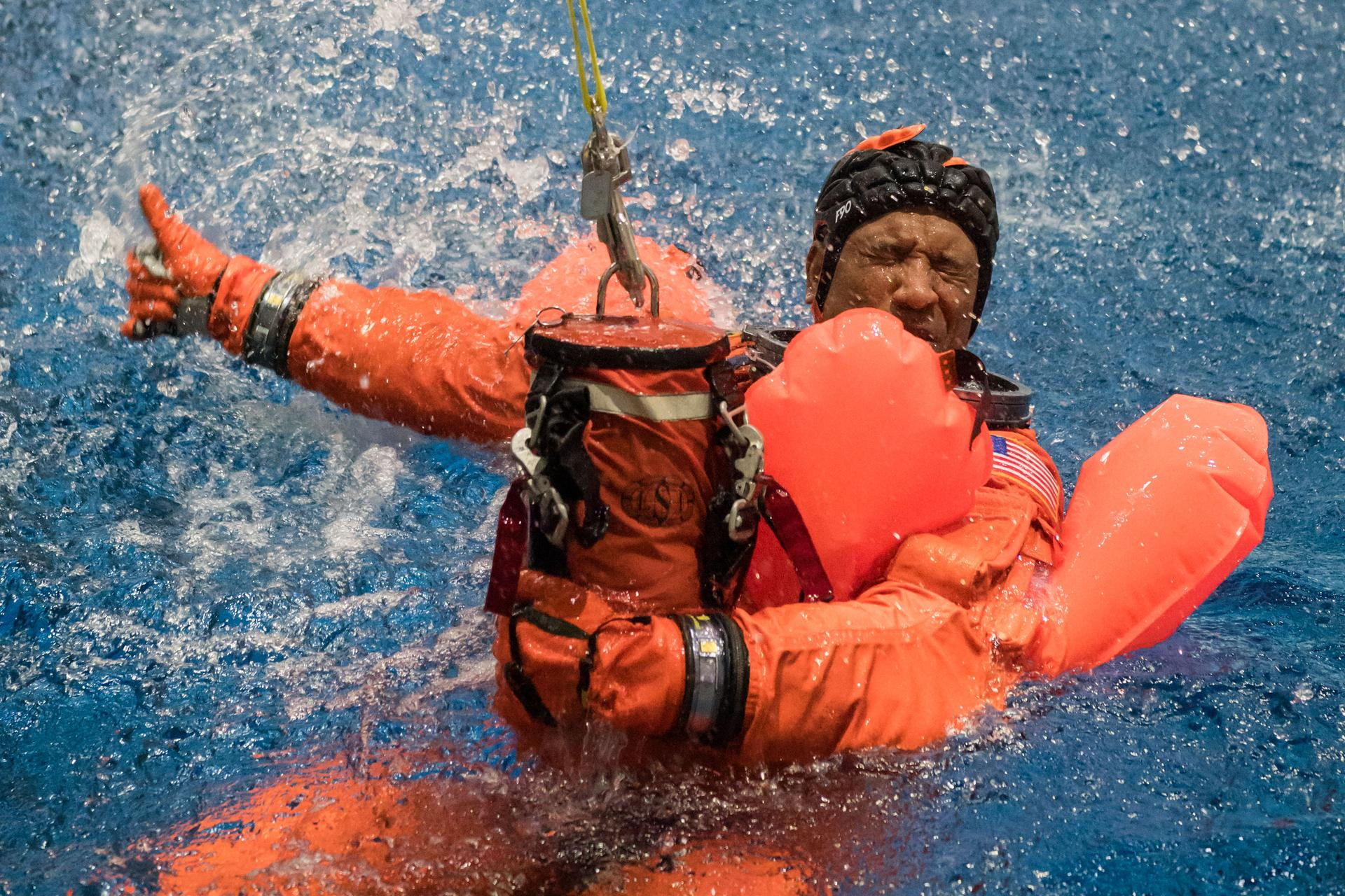 A man wearing a bright orange spacesuit and a bike helmet floats with on arm wrapped holding an orange sack, wrapped around an orange inflatable. He is half submerged in vibrant blue water, splashing his orange hand from the wet to give a thumbs up.