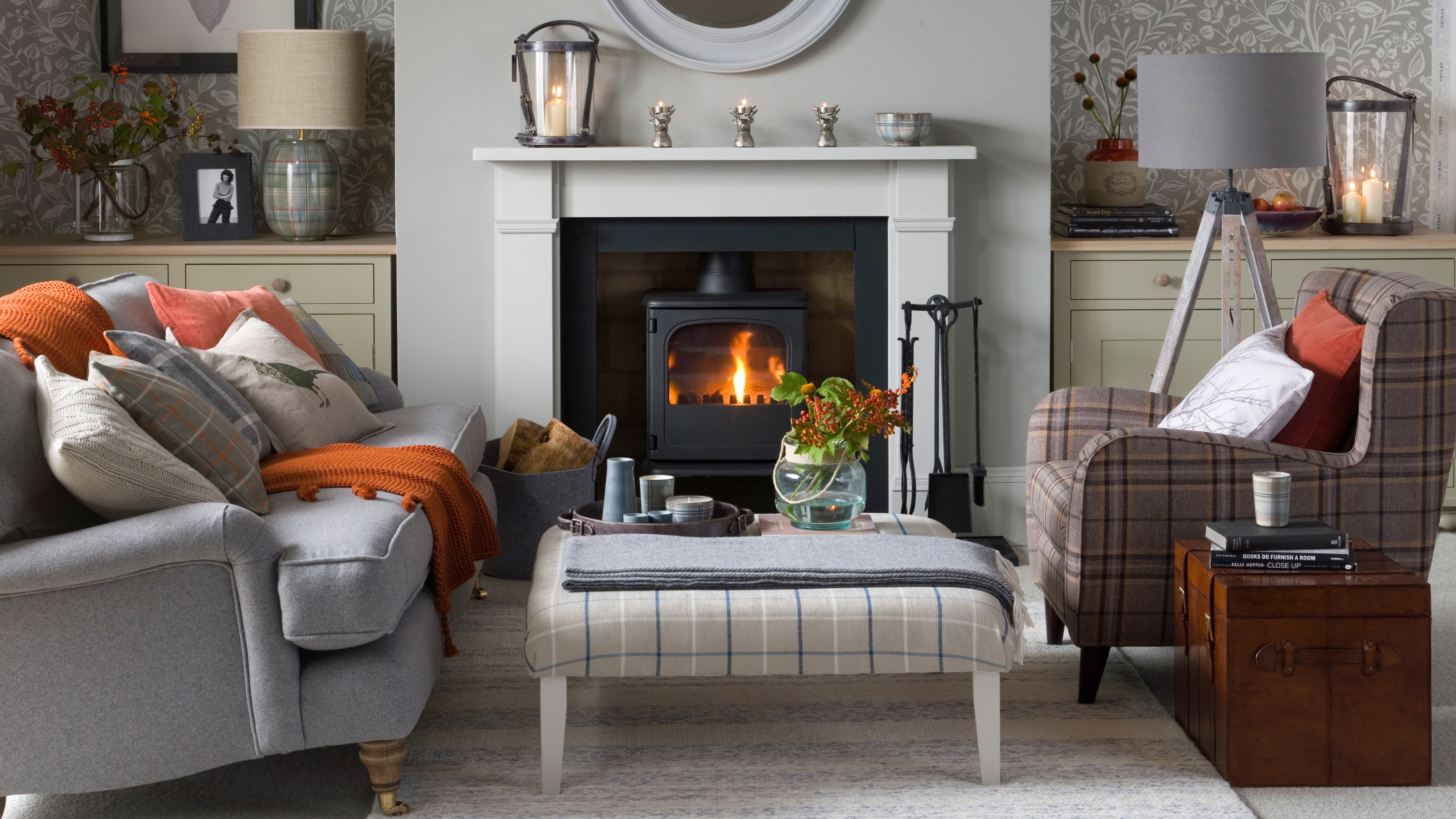 Grey modern country living room, nature pattern wallpaper, wood burning fireplace, lit fire, grey mantelpiece, grey sofa, plaid stool and armchair.