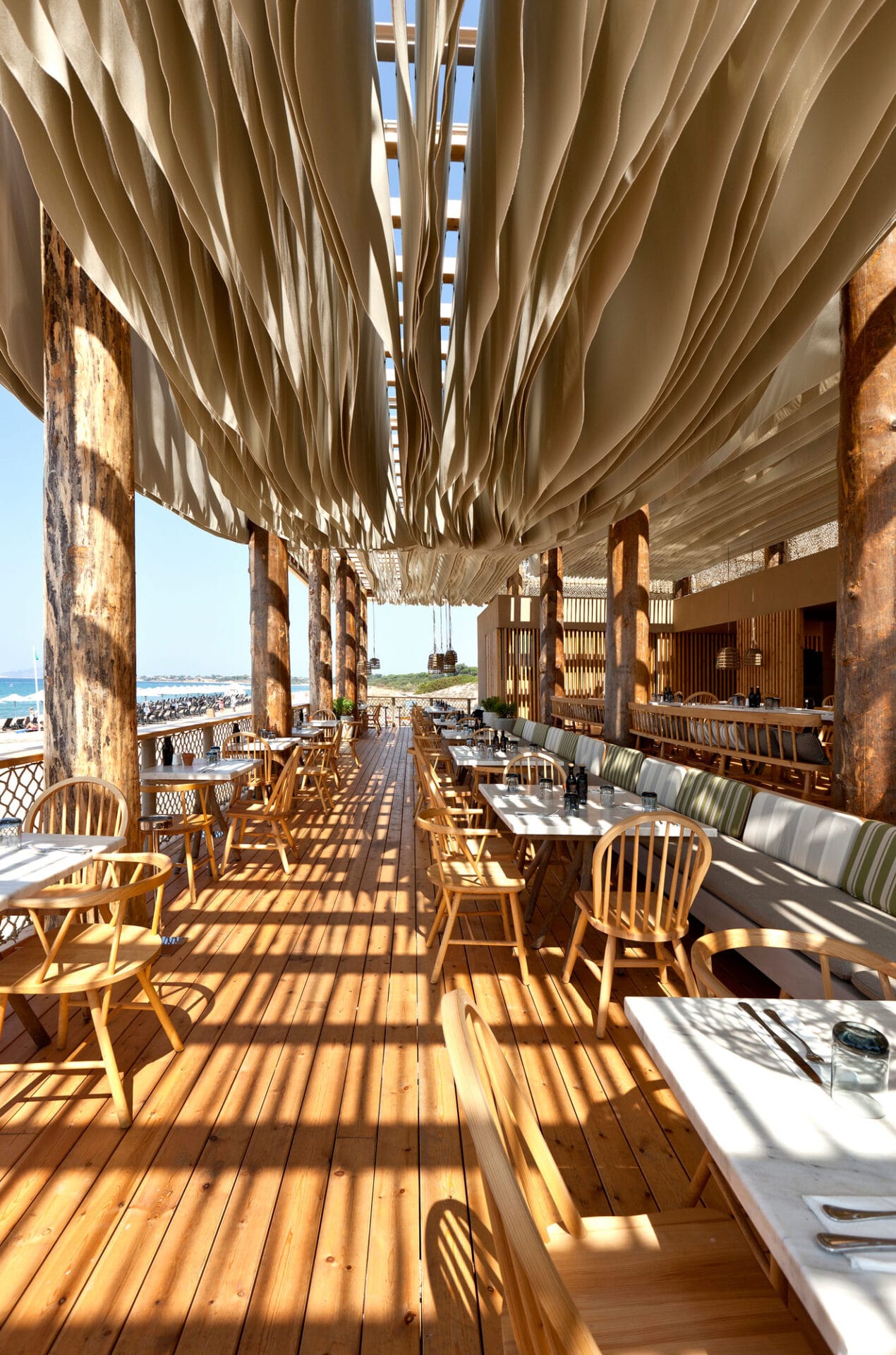A sea-view restaurant terrace with wooden floors and chairs, white stone tables, a stripy banquette in green and grey tones, and a terrace-wide ceiling installation made of multiple paper-like layers.