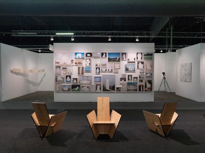 An installation view of three wooden chairs in the foreground and a wall with photos in frames 
