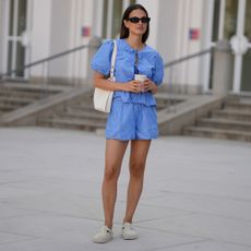 Anna Winter is seen wearing a blue white striped shorts and matching blue top from Rita Montezuma x NA-KD; a pair of off-white suede Boston slippers from Birkenstock; a light cream white small Romy hobo leather bag with a slim shoulder strap and silver button logo detail from Tory Burch; black rectangular Teru sunglasses from Ray-Ban on June 15, 2025 in Munich, Germany.