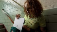 A woman with curly hair is shown sitting on a tiled staircase, carefully placing a slim, silver laptop into a green corduroy backpack.
