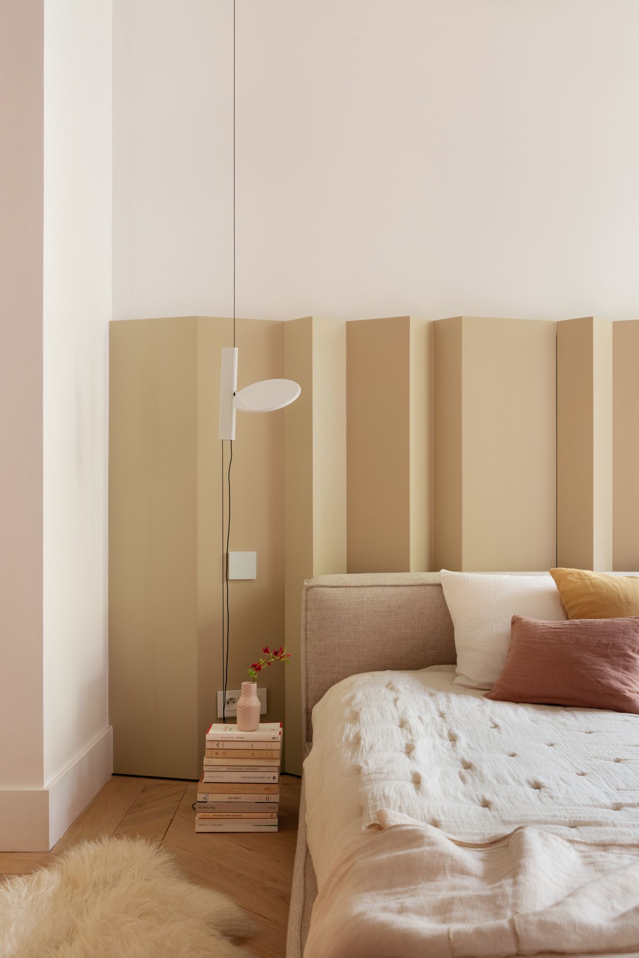 a grown up children's room with a large bed with a room divider design behind the headboard within a neutral color scheme; there are a pile of books next to the bed being used as a makeshift nightstand and an understated light hung from the ceiling