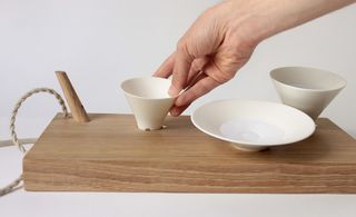 A hand adding a small white cermaic dish to a wood board holding 2 other white ceramic dishes. photographed against a white background