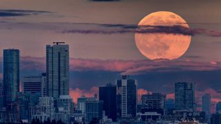 Large pink supermoon rises through wispy clouds above Vancouver city skyline. 