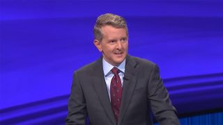 Ken Jennings looking serious at the podium on Jeopardy in a grey suit and red tie, Tournament of Champions.