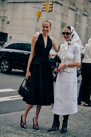 Two women wearing a black dress with a black bag and a white top and skirt with a silk scarf