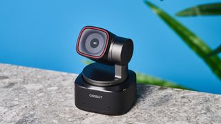 a small PTZ webcam with 4K resolution and a magnetic attachment clip is photographed in front of a blue background and a green plant