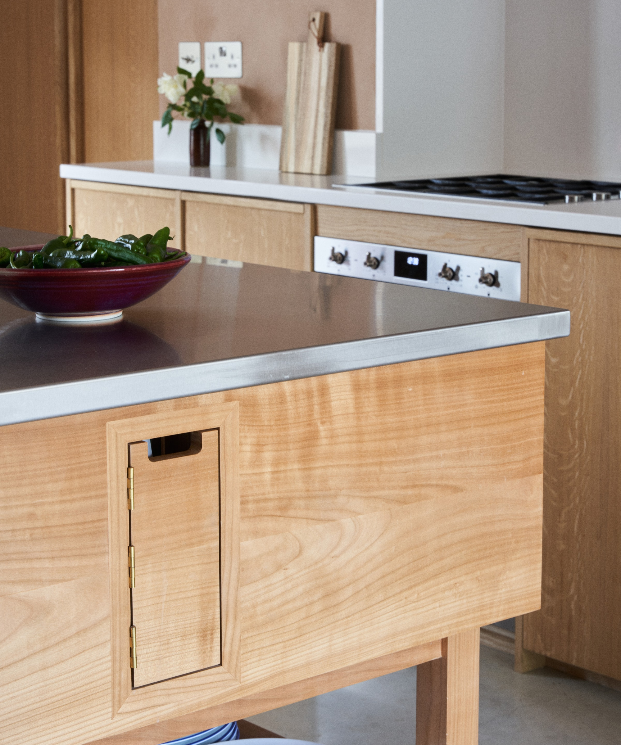 A wooden kitchen island hides a small hinged door built seamlessly into the panelled side. The stainless steel worktop above holds a bowl of fresh vegetables, while warm timber cabinetry creates a soft, natural aesthetic throughout the kitchen.