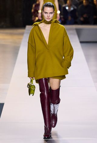 a photo showing some of the best thigh-high boots with a model walking the Gucci runway show wearing a mustard yellow deep v-neck mini dress styled with burgundy red tall boots with a green mini bag