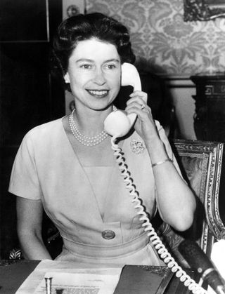 A black and white photo of Queen Elizabeth speaking on a corded telephone wearing pearls and a dress