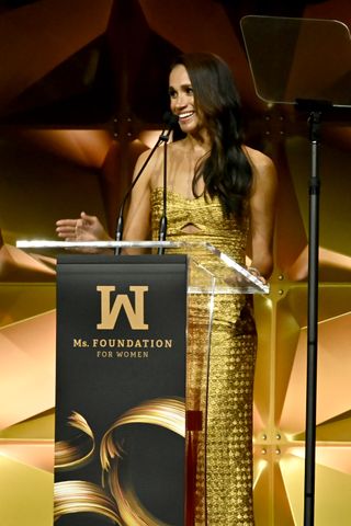 Meghan Markle wears a strapless gold brocade dress as she stands at a podium giving a speech