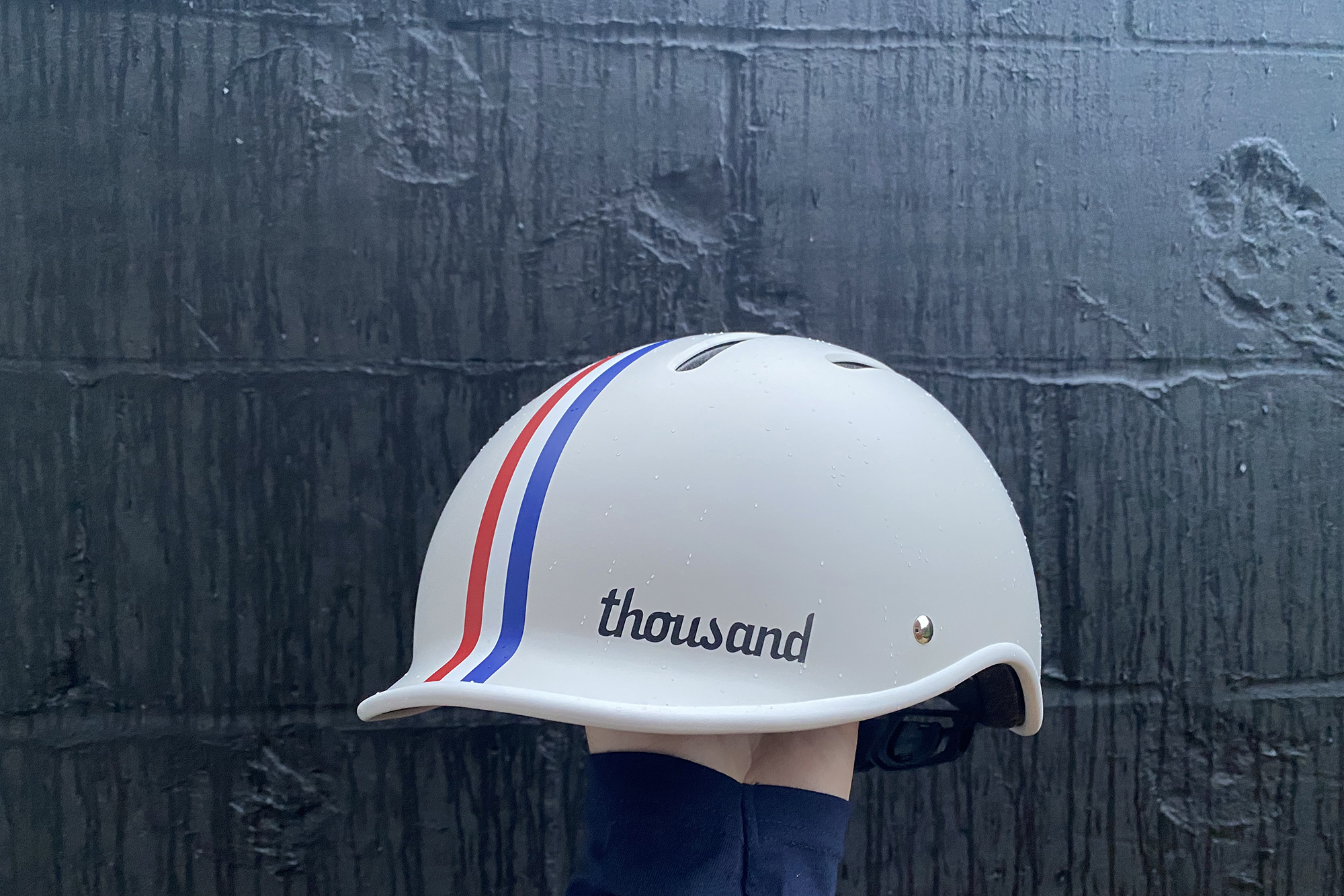 Thousand Jr kids helmet being held up against a wall painted black, in the White, with red,white and blue strip version.