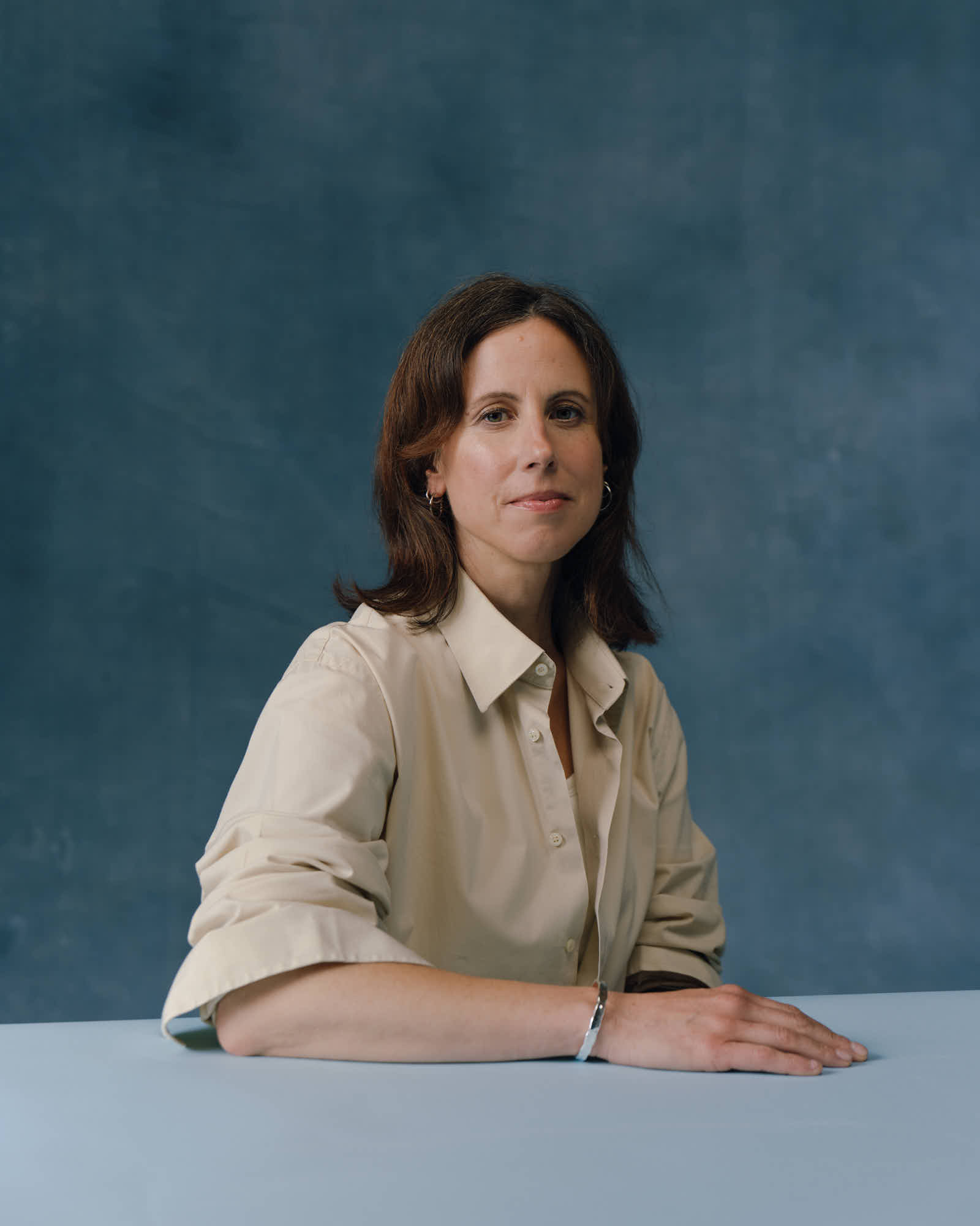 A woman in a cream shirt sits at a light blue table against a dark teal backdrop with one arm resting on the table and other below