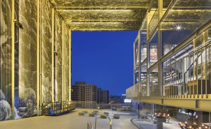 View of the Hyundai Music Library at night - a glass front building with a geometric design, outdoor seating area and wall in front with images of people