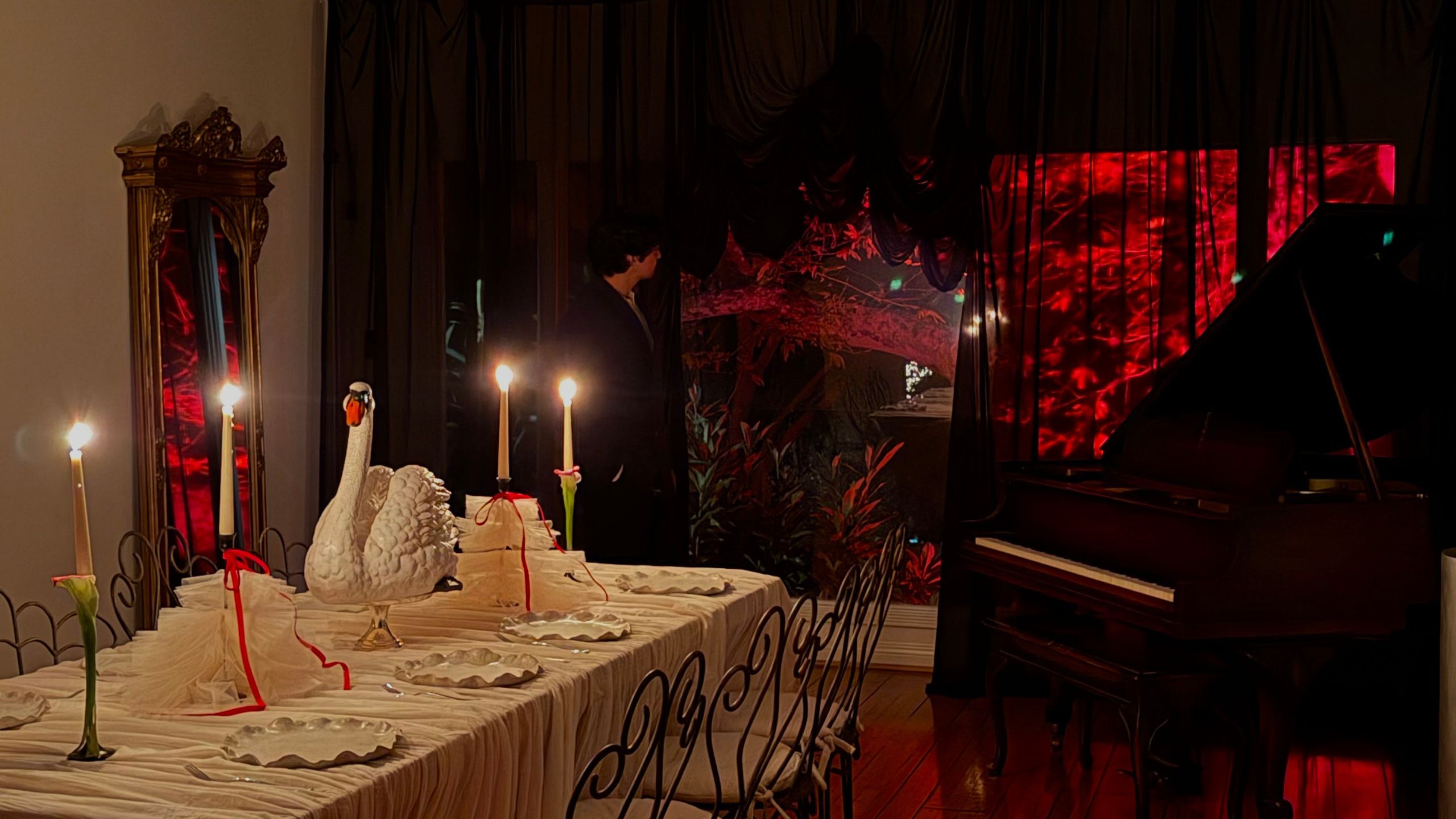 A dining area decorated for Halloween with black sheer curtains partly revealing a red-lit trees outdoors, a grand piano, and a table set with a swan statue in the center among taper candles