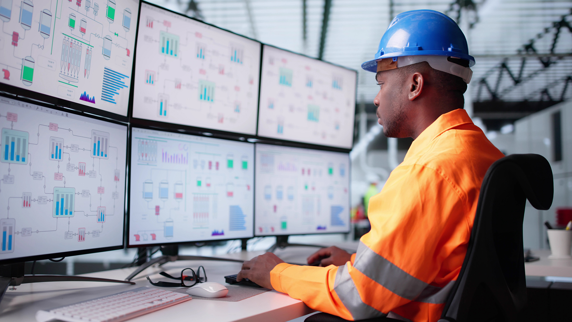 A site worker in a high-vis orange jacket and a blue hard hat watching six monitors displaying data from operational technology.