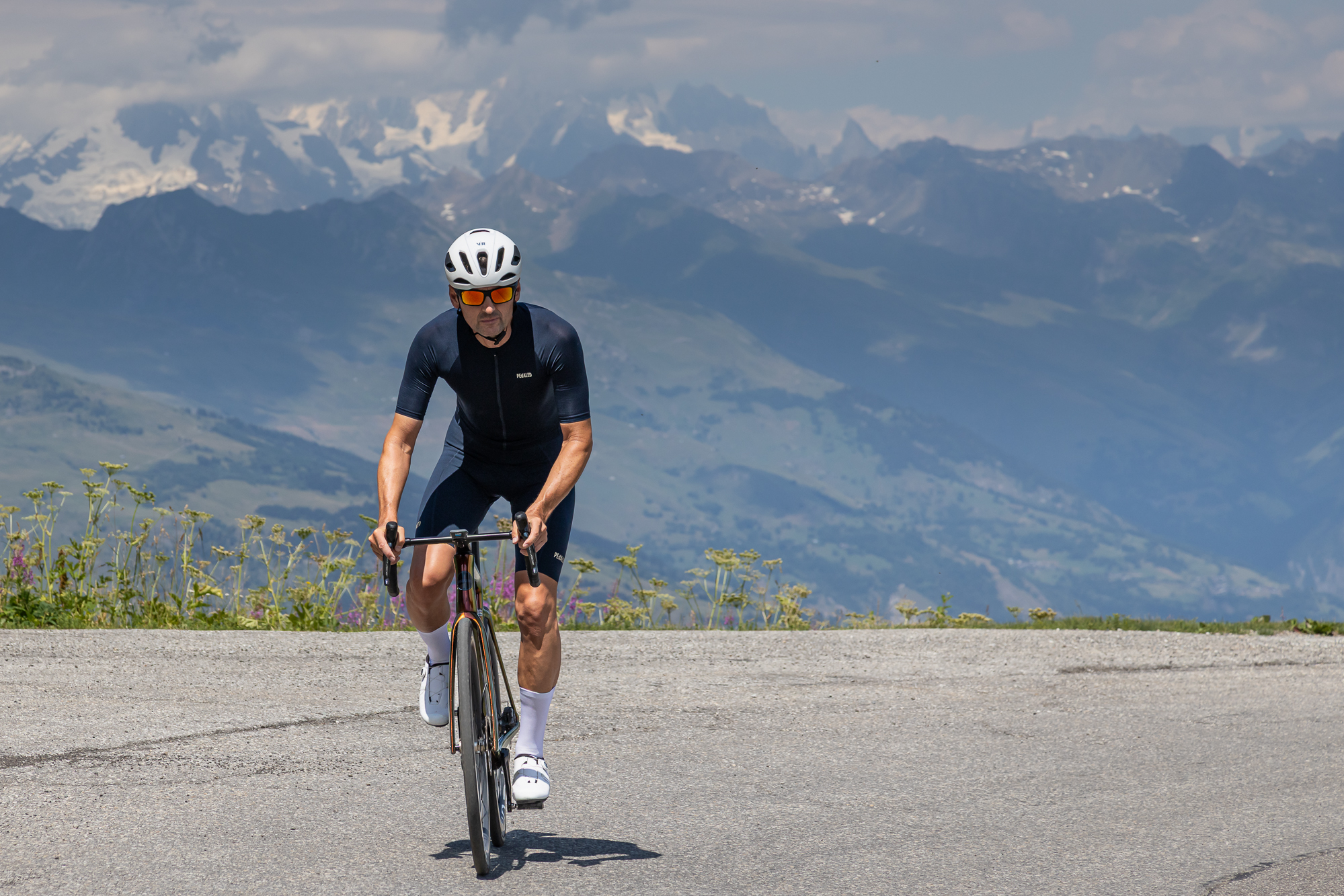 front view of a man riding an orange bike wearing a blue jersey, blue shorts, white helmet and white shoes with alpine mountains in the background
