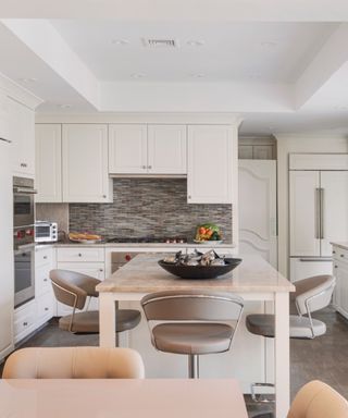 kitchen with off white cabinetry, kitchen island, taupe floor tiles and backsplash, taupe countertop, bar stools