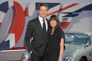 Kris Thykier and wife Claudia Winkleman attend the World Premiere of "No Time To Die" at the Royal Albert Hall wearing black suit and a black dress against a Union Jack background