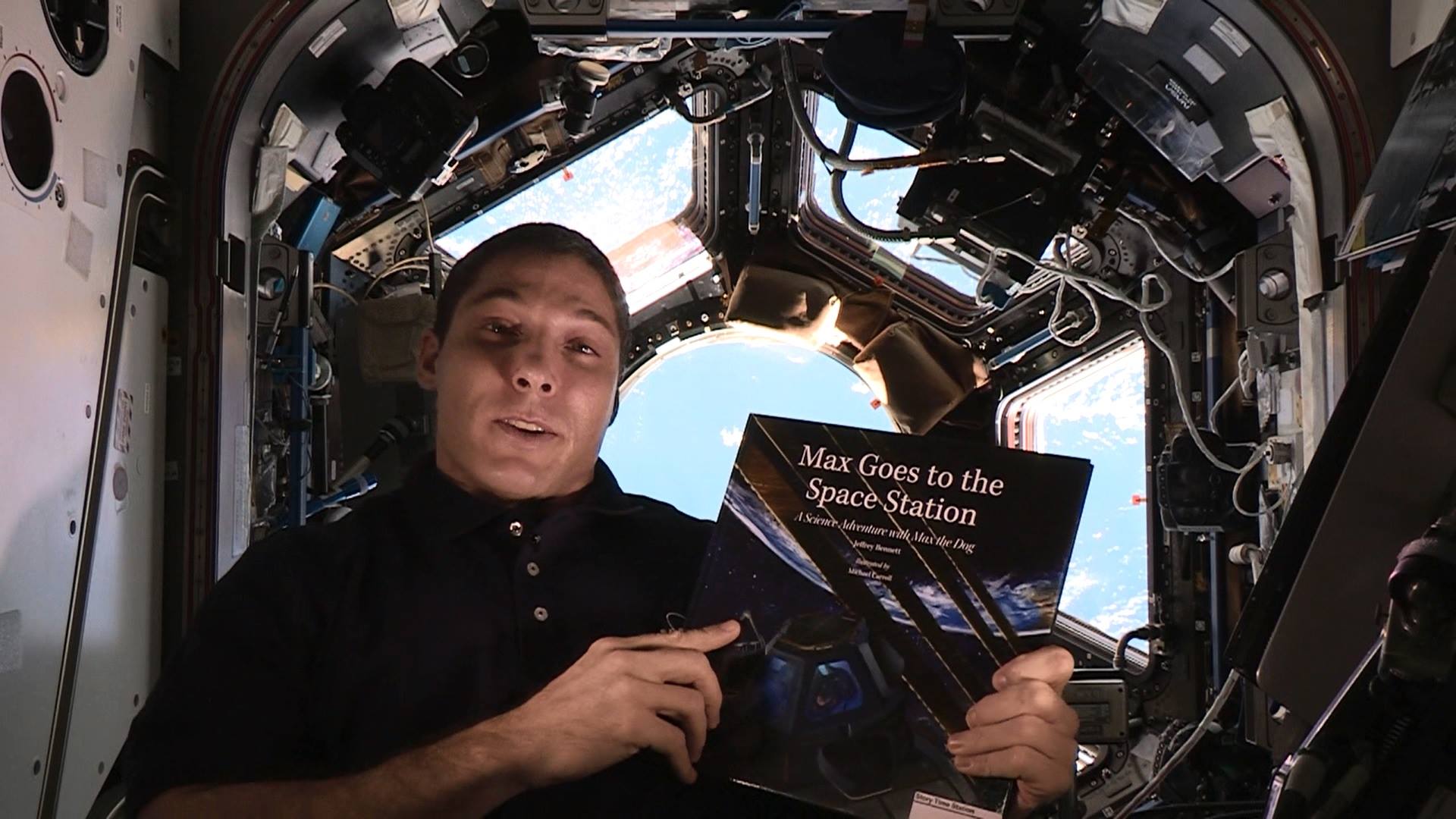 NASA astronaut Mike Hopkins holds the book &quot;Max Goes to the Space Station&quot; inside the real-life space station&#039;s Cupola observation deck. The book, written by author Jeffrey Bennett, is part of Story Time From Space.