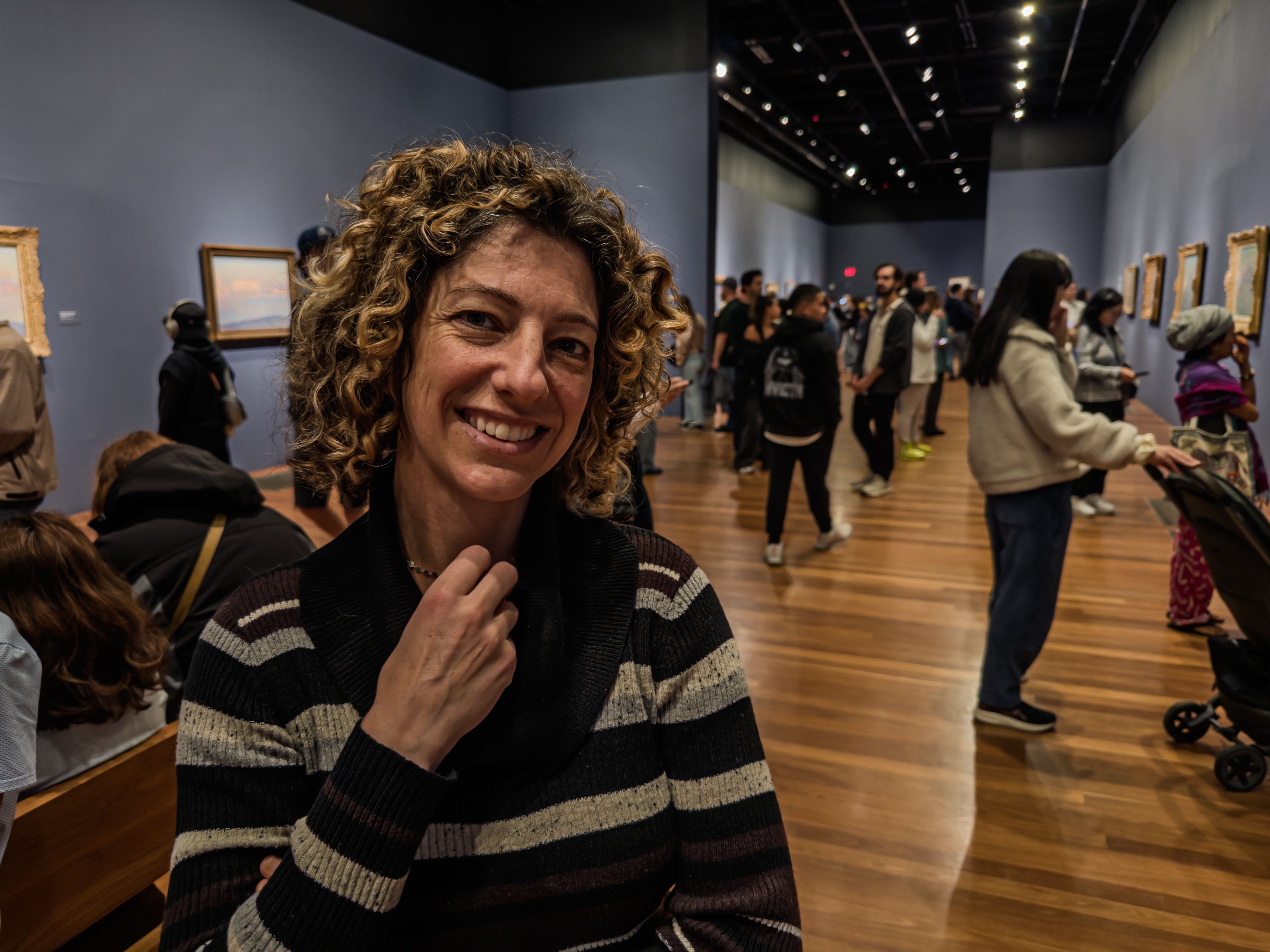 Smiling woman with curly hair poses in a busy art gallery filled with visitors and framed paintings.