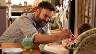 Taran Killam feeding a baby with a spoon while leaning over a kitchen table.