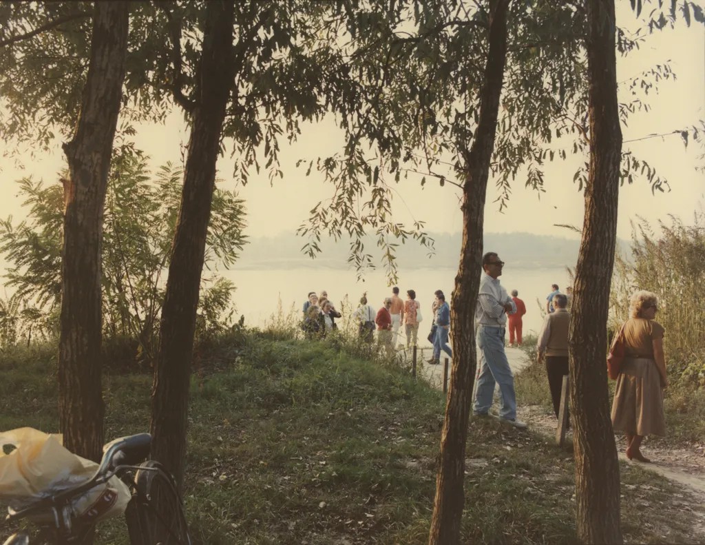A lake view populated by a group of people dressed in colorful clothing features multiple trees, lots of greenery, and a vintage bike positioned in its left-hand corner.