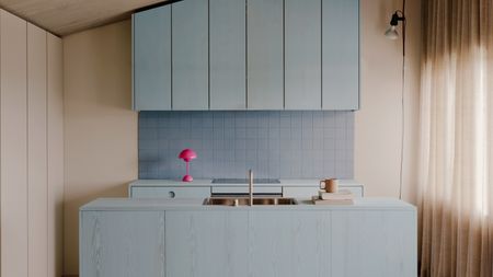 Image of a minimalist kitchen design. The cabinets are stained in a light blue color over natural wood. The backsplash is also a light blue tile, and there is a hot pink, mini mushroom lamp on the counter.